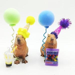May include: Two capybara figurines wearing party hats and holding balloons. One capybara is holding a green balloon and the other is holding a blue balloon. Both capybaras are wearing party hats with colourful decorations. The capybaras are standing on a white surface. The capybara on the left is holding a small glass of a milkshake with a straw. The capybara on the right is holding a small bag of sweets with the text "Haribo Pinkie & Lilly" printed on it.