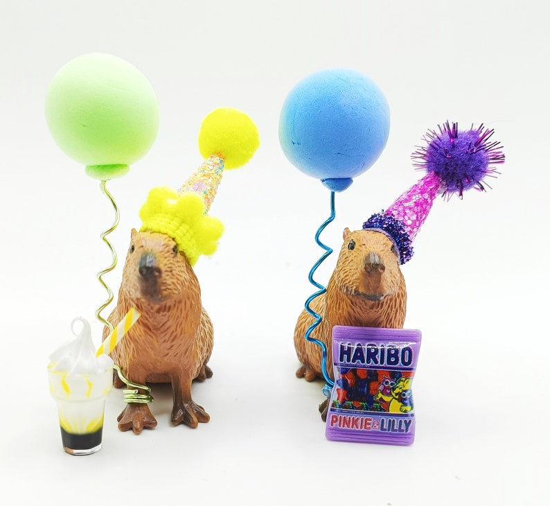 May include: Two capybara figurines wearing party hats and holding balloons. One capybara is holding a green balloon and the other is holding a blue balloon. Both capybaras are wearing party hats with colourful decorations. The capybaras are standing on a white surface. The capybara on the left is holding a small glass of a milkshake with a straw. The capybara on the right is holding a small bag of sweets with the text "Haribo Pinkie & Lilly" printed on it.