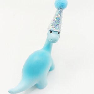 May include: A light blue dinosaur figurine wearing a silver and blue party hat with a blue pom-pom on top.