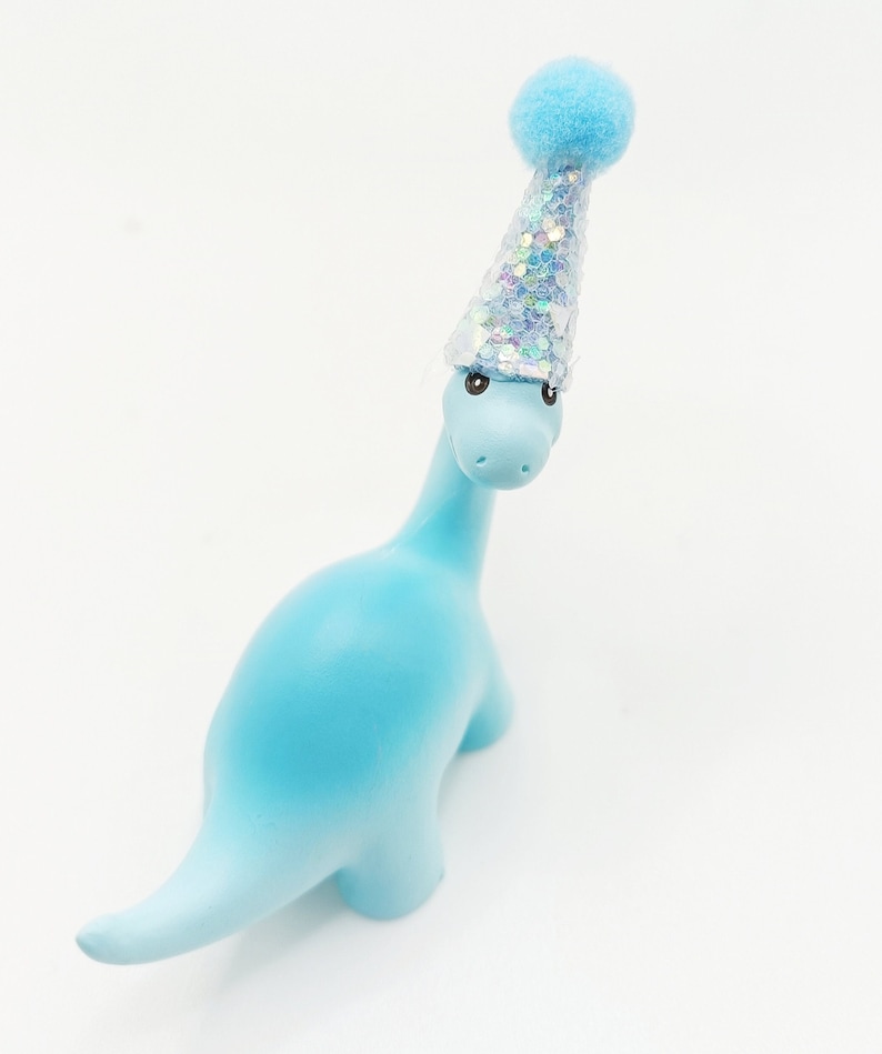 May include: A light blue dinosaur figurine wearing a silver and blue party hat with a blue pom-pom on top.