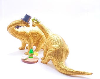 May include: Two gold dinosaur figurines, one wearing a top hat and the other a flower crown, stand on a wooden base with a bottle of champagne and two glasses.