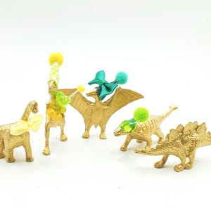 Puede incluir: Cinco figuritas de dinosaurio doradas con decoraciones coloridas. Los dinosaurios son un estegosaurio, un pterod&aacute;ctilo, un braquiosaurio y dos triceratops. Las figuritas est&aacute;n decoradas con pompones y cintas verdes, azules y amarillas.