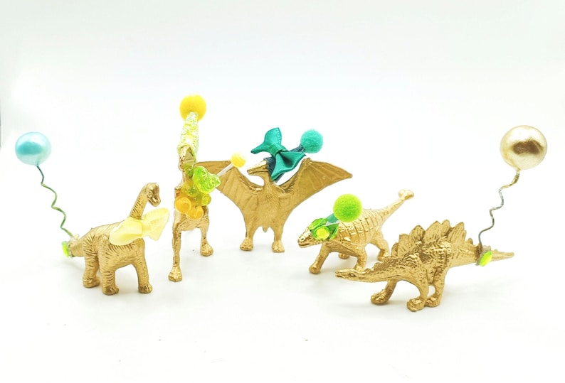 Puede incluir: Cinco figuritas de dinosaurio doradas con decoraciones coloridas. Los dinosaurios son un estegosaurio, un pterod&aacute;ctilo, un braquiosaurio y dos triceratops. Las figuritas est&aacute;n decoradas con pompones y cintas verdes, azules y amarillas.