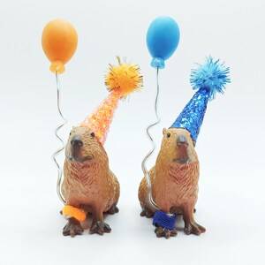 May include: Two capybaras wearing party hats and holding balloons. One capybara is wearing a blue hat and holding a blue balloon, the other is wearing an orange hat and holding an orange balloon.