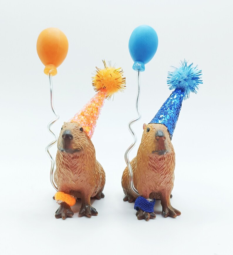 May include: Two capybaras wearing party hats and holding balloons. One capybara is wearing a blue hat and holding a blue balloon, the other is wearing an orange hat and holding an orange balloon.
