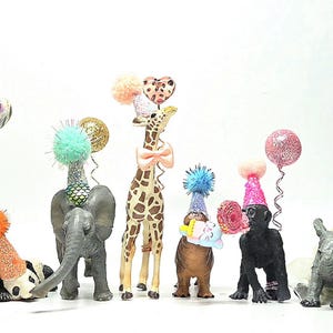 May include: A collection of animal figurines, including a zebra, panda, elephant, giraffe, and gorilla, each adorned with party hats and decorative balloons. The figurines are in various colors and poses, creating a festive scene.