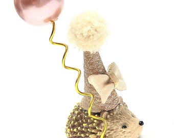 Può includere: Una piccola statuetta di riccio che indossa un cappellino da festa dorato e glitterato, sormontato da un pon-pon color crema e un piccolo fiocco. Un filo dorato parte dalla base e sostiene un palloncino rosa metallizzato, rotondo e lucido.