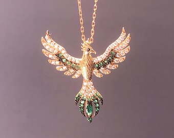 May include: Gold-coloured phoenix pendant necklace with a detailed design. The phoenix has outstretched wings adorned with clear and emerald-green gemstones. The necklace chain is also gold-coloured, creating an elegant piece of jewellery.