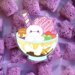 May include: A clear acrylic sticker featuring a cartoon dessert bowl with a white character, ice cream, and fruit. The bowl has a yellow and blue rim, with the text "PINNY PLANET". A purple and white striped stick is on the left.