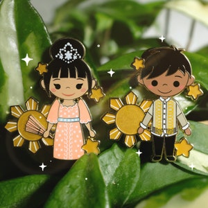 Puede incluir: Dos pines de esmalte con figuras de dibujos animados con vestimenta tradicional filipina. La figura femenina lleva un vestido melocotón y una tiara, sosteniendo un abanico. La figura masculina viste un atuendo beige y amarillo. Ambos pines tienen un diseño de sol dorado.