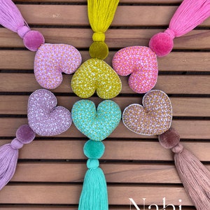 May include: Six heart-shaped fabric ornaments with floral patterns in various colors, each attached to a tassel. The tassels are in pink, yellow, purple, green, and brown.