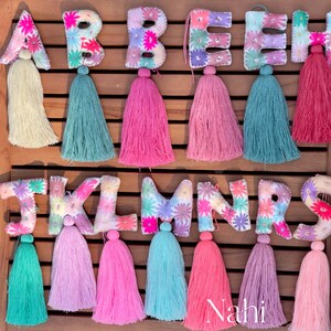 May include: A set of felt letter charms with colorful flower embroidery and tassel accents. The letters are A, B, C, D, E, F, G, H, I, J, K, L, M, N, O, P, Q, R, S, and T. The tassels are in various colors, including pink, green, and purple.