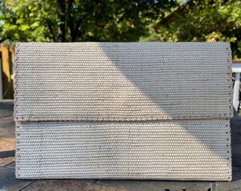 May include: A beige woven clutch purse with a pink stitched border. The purse is closed and sitting on a brown tiled surface.