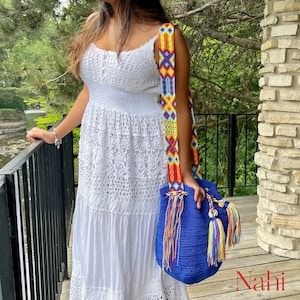 May include: A woman wearing a white dress with a blue and multi-colored crocheted bag with tassels. The bag has a long, colorful strap. The bag is hanging from her shoulder. The word "Nahi" is written in red on the bottom right corner of the image.