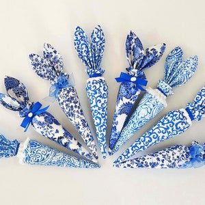 K&ouml;nnte beinhalten: Neun blaue und wei&szlig;e Stoff-Osterhasen-Karotten mit blauen Schleifen. Die Karotten sind kegelf&ouml;rmig und haben ein blaues und wei&szlig;es Blumenmuster.
