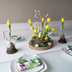 Könnte beinhalten: Ein weißer Tisch mit einem Mittelstück aus gelben Tulpen, Weidenzweigen und goldenen Eiern in einem geflochtenen Nest. Der Tisch ist mit weißen Tischsets, weißen Tellern und grün-weiß gemusterten Servietten gedeckt.