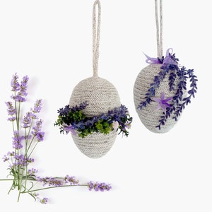 Puede incluir: Dos adornos blancos, tejidos, en forma de huevo con ramitas de lavanda y una corona verde. Los adornos cuelgan de un fondo blanco.
