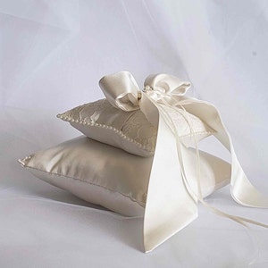 May include: A white satin ring pillow with a lace overlay and a white satin bow. The pillow is decorated with pearls.