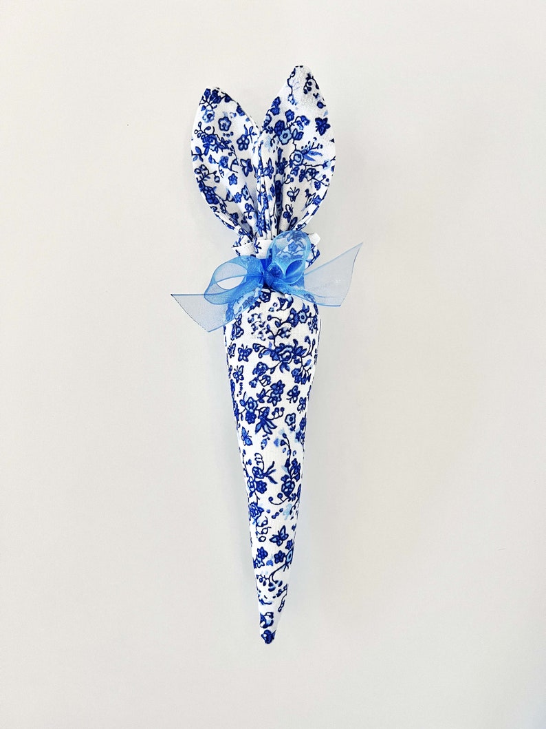 Peut inclure: Un c&ocirc;ne de P&acirc;ques en forme de lapin en tissu floral bleu et blanc avec un ruban bleu et des accents de perles blanches.