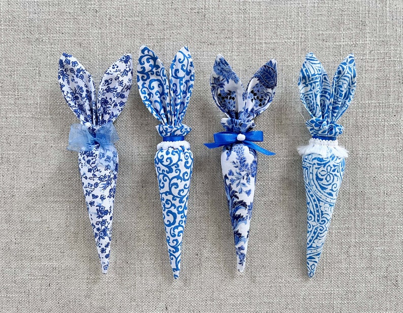 K&ouml;nnte beinhalten: Vier blaue und wei&szlig;e Stoff-Karotten in Hasenform mit blauen Schleifenakzenten. Die Karotten sind mit einem blauen und wei&szlig;en Blumenmuster verziert.