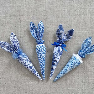 K&ouml;nnte beinhalten: Vier blaue und wei&szlig;e Stoff-Osterhasen-Karotten mit blauen Schleifen. Die Karotten liegen auf einem hellgrauen, strukturierten Hintergrund.