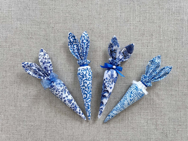 K&ouml;nnte beinhalten: Vier blaue und wei&szlig;e Stoff-Osterhasen-Karotten mit blauen Schleifen. Die Karotten liegen auf einem hellgrauen, strukturierten Hintergrund.