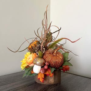 Puede incluir: Un centro de mesa de otoño con una calabaza naranja brillante, flores amarillas, piñas y un hongo en una maceta de madera.