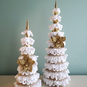 May include: Two white and gold Christmas trees with a gold flower accent. The trees are made of white yarn and have a gold base.
