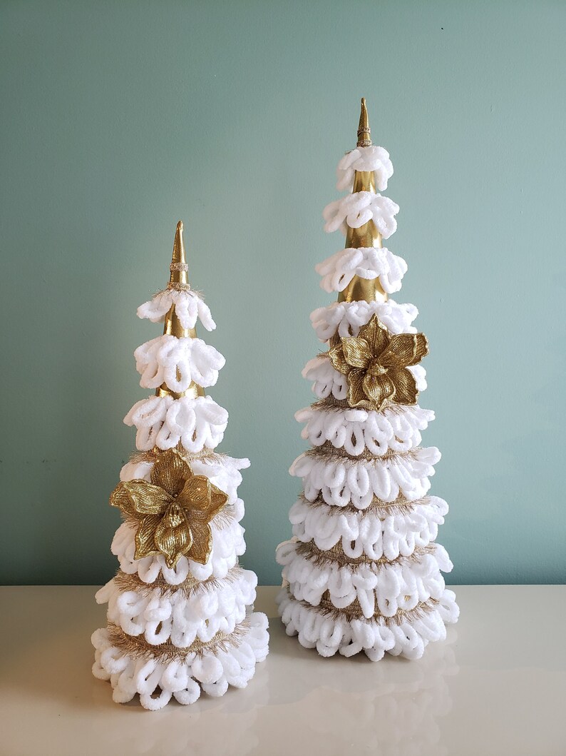 May include: Two white and gold Christmas trees with a gold flower accent. The trees are made of white yarn and have a gold base.