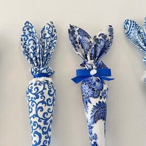 K&ouml;nnte beinhalten: Vier blaue und wei&szlig;e Stoff-Osterhasen-Kegel. Die Kegel sind mit einem blauen und wei&szlig;en Blumenmuster verziert. Zwei der Kegel haben blaue Schleifen, einer hat einen wei&szlig;en Bommel und eine blaue Schleife und einer hat eine wei&szlig;e Spitzenborte.