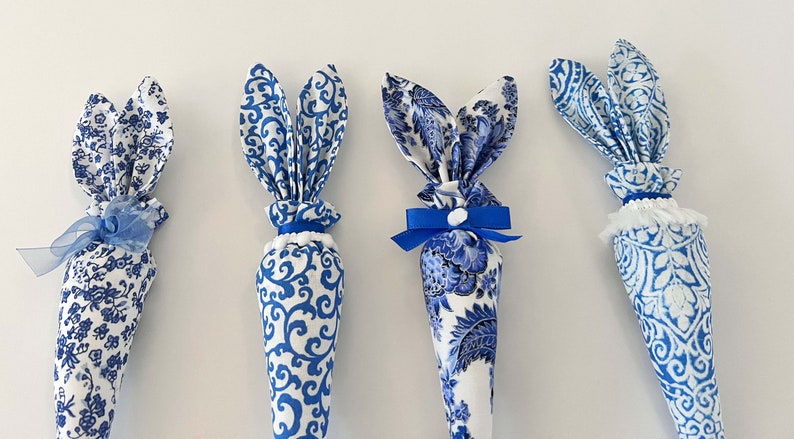 K&ouml;nnte beinhalten: Vier blaue und wei&szlig;e Stoff-Osterhasen-Kegel. Die Kegel sind mit einem blauen und wei&szlig;en Blumenmuster verziert. Zwei der Kegel haben blaue Schleifen, einer hat einen wei&szlig;en Bommel und eine blaue Schleife und einer hat eine wei&szlig;e Spitzenborte.