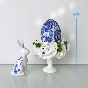 Peut inclure: Un pi&eacute;destal en c&eacute;ramique blanche surmont&eacute; d'un &oelig;uf de P&acirc;ques &agrave; motifs bleu et blanc et de fleurs blanches. Le pi&eacute;destal mesure 23 cm de haut. Un lapin en c&eacute;ramique blanche avec des accents floraux bleus se trouve au premier plan.