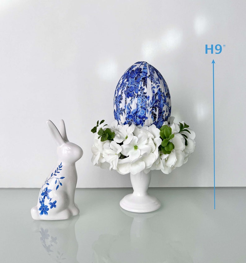 Peut inclure: Un pi&eacute;destal en c&eacute;ramique blanche surmont&eacute; d'un &oelig;uf de P&acirc;ques &agrave; motifs bleu et blanc et de fleurs blanches. Le pi&eacute;destal mesure 23 cm de haut. Un lapin en c&eacute;ramique blanche avec des accents floraux bleus se trouve au premier plan.