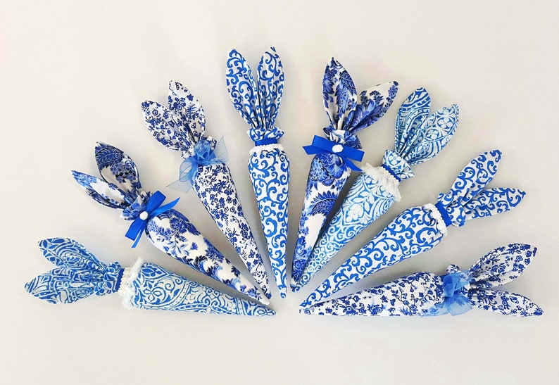 K&ouml;nnte beinhalten: Acht blaue und wei&szlig;e Stoff-Hasenkegel mit blauen Schleifen. Die Kegel sind mit einem blauen und wei&szlig;en Blumenmuster verziert.