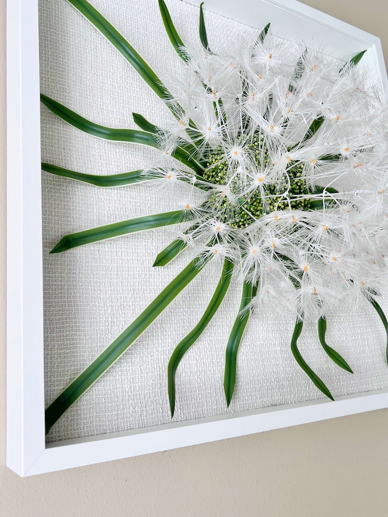 May include: A framed wall art piece featuring a white dandelion with green leaves on a white textured background.
