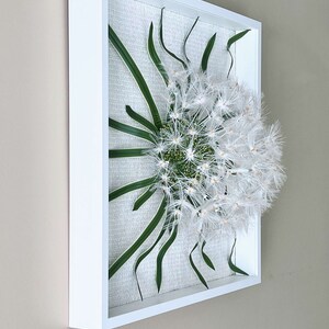May include: A square wall art piece with a white frame, featuring a white dandelion with green leaves on a textured white background.