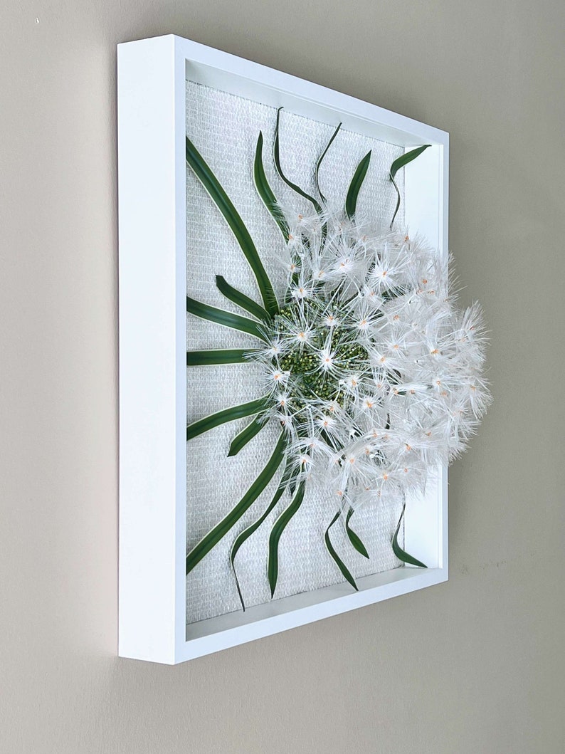 May include: A square wall art piece with a white frame, featuring a white dandelion with green leaves on a textured white background.