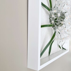 May include: A square wall art piece with a white dandelion and green leaves inside. The dandelion is in the centre of the frame and the leaves are spread out around it.