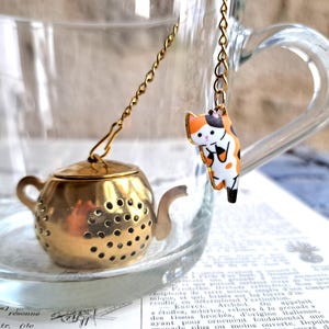 Pode incluir: Infusor de chá em metal dourado em forma de bule com tampa e corrente a condizer, pendurado dentro de uma chávena de chá de vidro transparente. Um pequeno pingente de gato colorido está preso à corrente.
