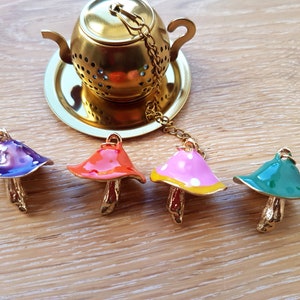 Puede incluir: Cuatro infusores de té con forma de hongo de colores con cadenas doradas. Los infusores son morado, rojo, rosa con puntos blancos y verde. Los infusores están sobre una superficie de madera junto a un colador de té dorado.