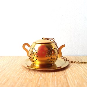 Peut inclure: Infuseur &agrave; th&eacute; en forme de th&eacute;i&egrave;re dor&eacute;e avec un couvercle et une cha&icirc;ne. L'infuseur repose sur une soucoupe dor&eacute;e assortie.
