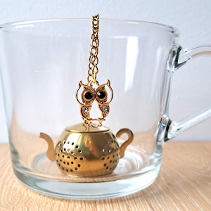 Może przedstawiać: Złoty zaparzacz do herbaty w kształcie czajnika z zawieszką w kształcie złotej sowy przymocowaną do pokrywki. Zaparzacz znajduje się w przezroczystej szklanej filiżance.
