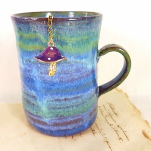 Könnte beinhalten: Eine blaue und grüne Keramiktasse mit einer goldenen Kette und einem violetten Pilz-Anhänger, der am Rand hängt.