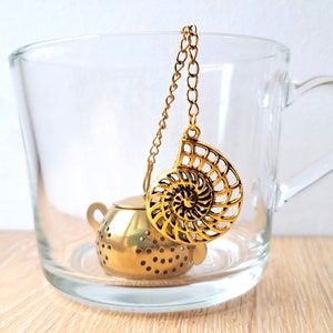 Puede incluir: Una taza de vidrio transparente contiene un infusor de té dorado. El infusor tiene un diseño de concha y una pequeña cesta en forma de tetera con perforaciones. Una cadena dorada conecta el infusor con la parte superior de la taza. La taza está sobre una superficie de madera clara.