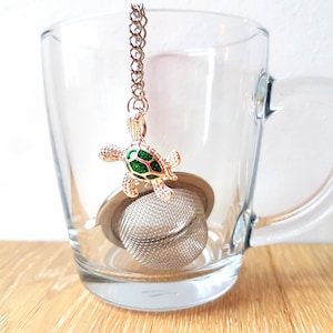 Può includere: Un infusore per tè in metallo con un cestello a maglia fine e una catenella color argento attaccata a un ciondolo decorativo a forma di tartaruga. Il ciondolo presenta una cornice in metallo dorato con un inserto glitterato verde sul guscio.