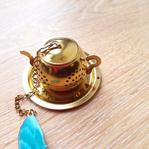 Peut inclure: Infuseur &agrave; th&eacute; en forme de th&eacute;i&egrave;re dor&eacute;e avec une cha&icirc;ne et un pendentif en pierre pr&eacute;cieuse bleu sarcelle. L'infuseur a un couvercle et une poign&eacute;e.