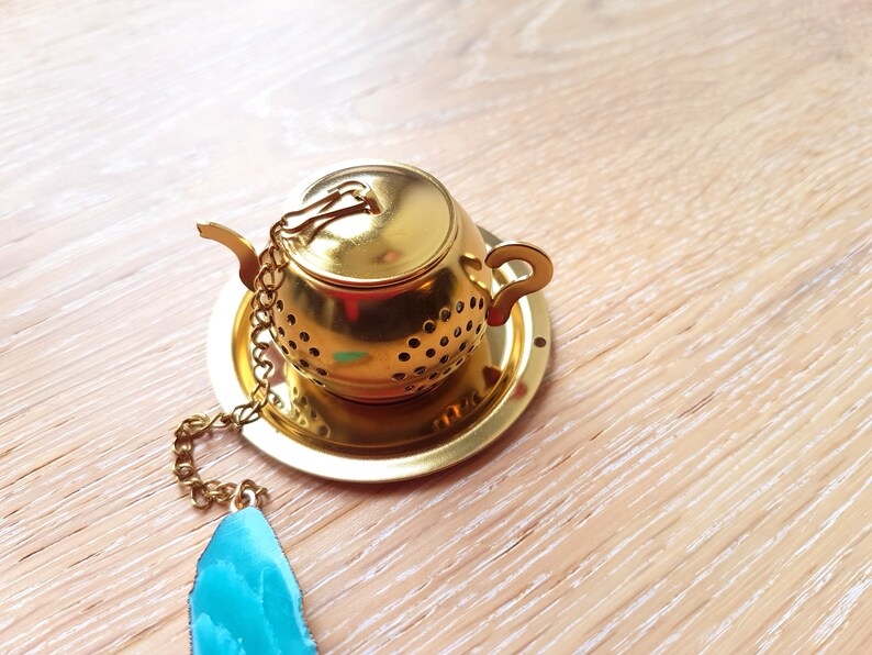 Peut inclure: Infuseur &agrave; th&eacute; en forme de th&eacute;i&egrave;re dor&eacute;e avec une cha&icirc;ne et un pendentif en pierre pr&eacute;cieuse bleu sarcelle. L'infuseur a un couvercle et une poign&eacute;e.