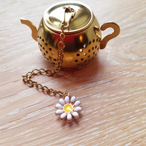 Könnte beinhalten: Goldener Teekanne-förmiger Tee-Ei mit einem weißen Gänseblümchen-Anhänger an der Kette. Der Tee-Ei hat einen Deckel und einen Boden aus Mesh.