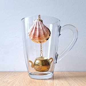 Può includere: Un infusore da tè dorato a forma di teiera con un coperchio a conchiglia appeso in una tazza di vetro trasparente con manico.