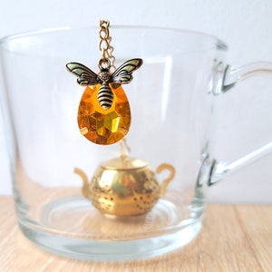 Könnte beinhalten: Ein goldener Tee-Ei in Form einer Teekanne mit einem Bienen- und Bernstein-Kristall-Anhänger. Der Tee-Ei befindet sich in einer durchsichtigen Glas Tasse.